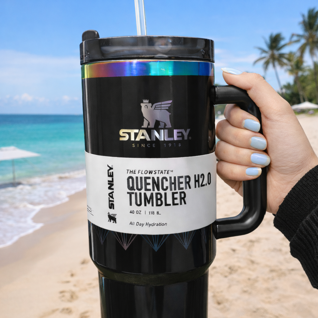Person holding a Stanley tumbler on a beach with ocean and palm trees in the background